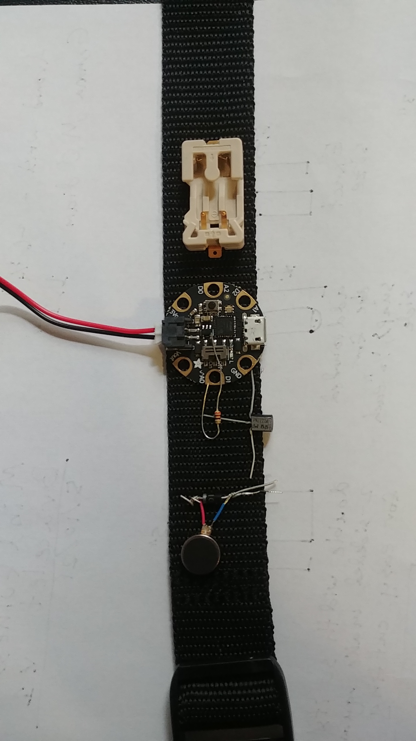black nylon strap with gemma on it. The strap is on a table going up and down the frame. below the gemma is the vibration circuit, laid out but not connected