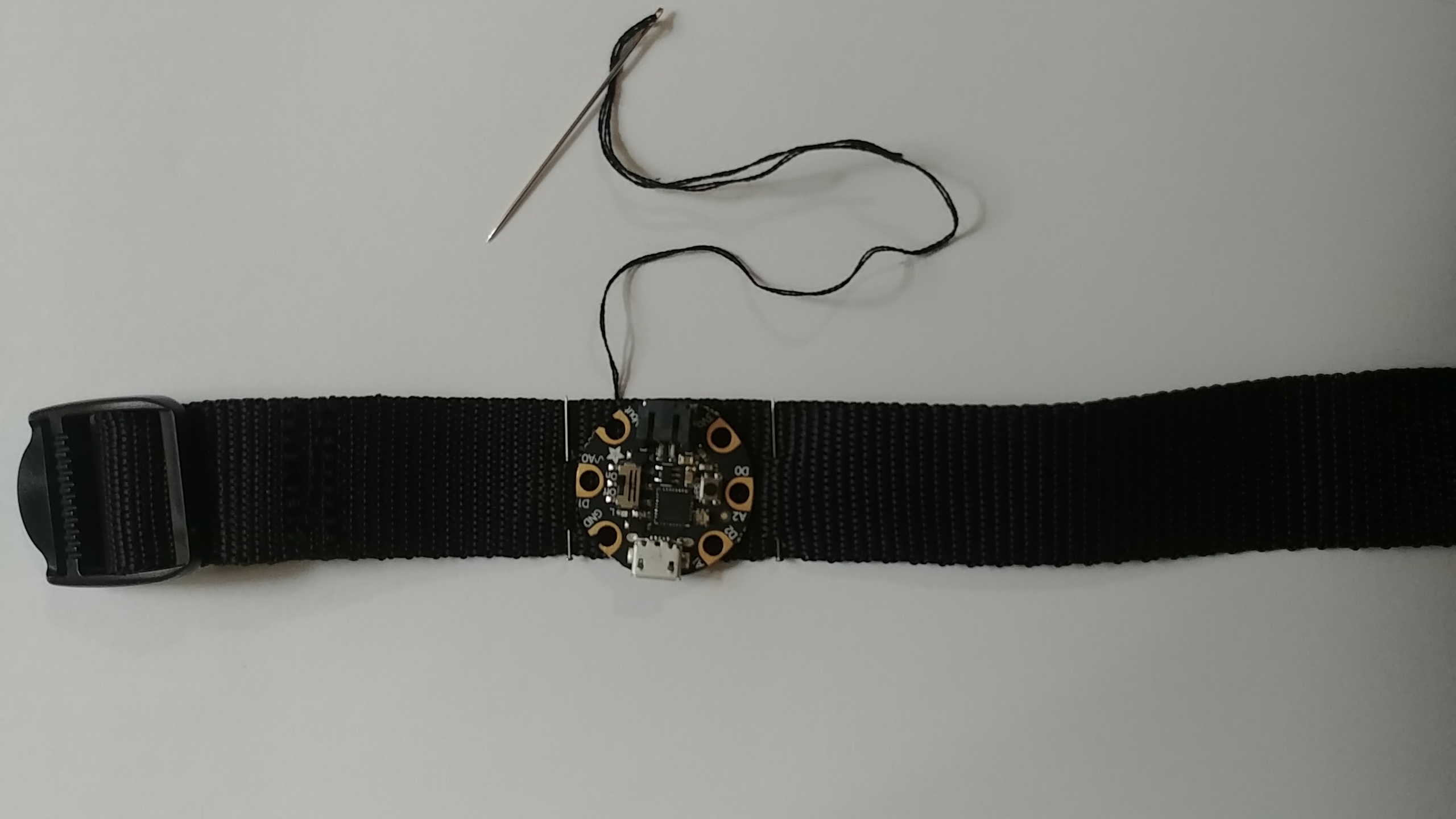 black nylon strap with the gemma on it, there is black thread coming from the bracelet, attached to a sewing needle. There are two sewing pins on either side of the gemma
