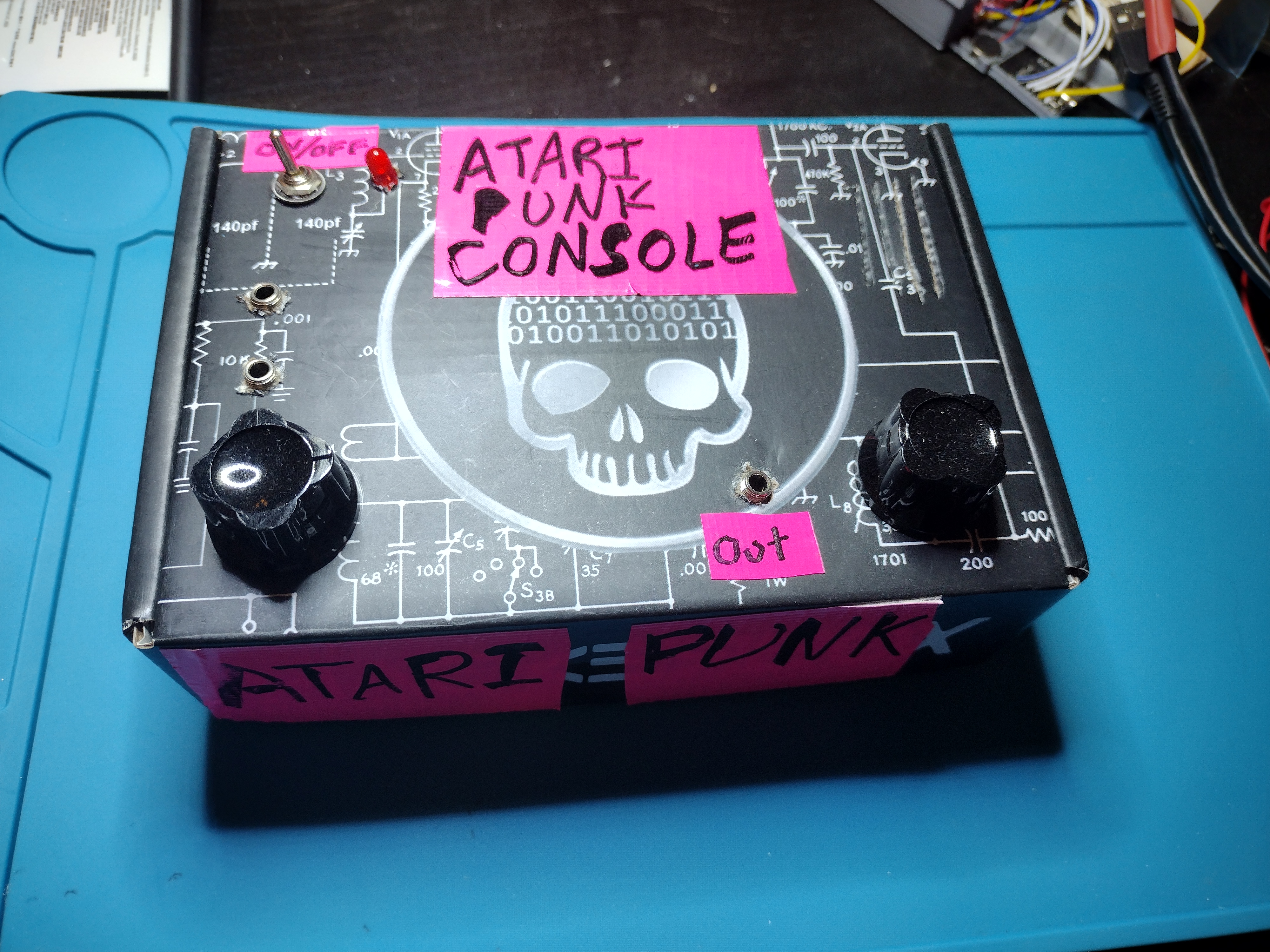 Picture of a black box on a blue anti-static mat. The box has a toggle switch and LED in the upper left corner, two knobs, one in each bottom left corner, and the words "Atari Punk Console" written in sharpie on pink duct tape taped to the top and side of the box. There are 3 headphone jacks sticking out the top of the box. One has the word "out" next to it in sharpie on pink duct tape