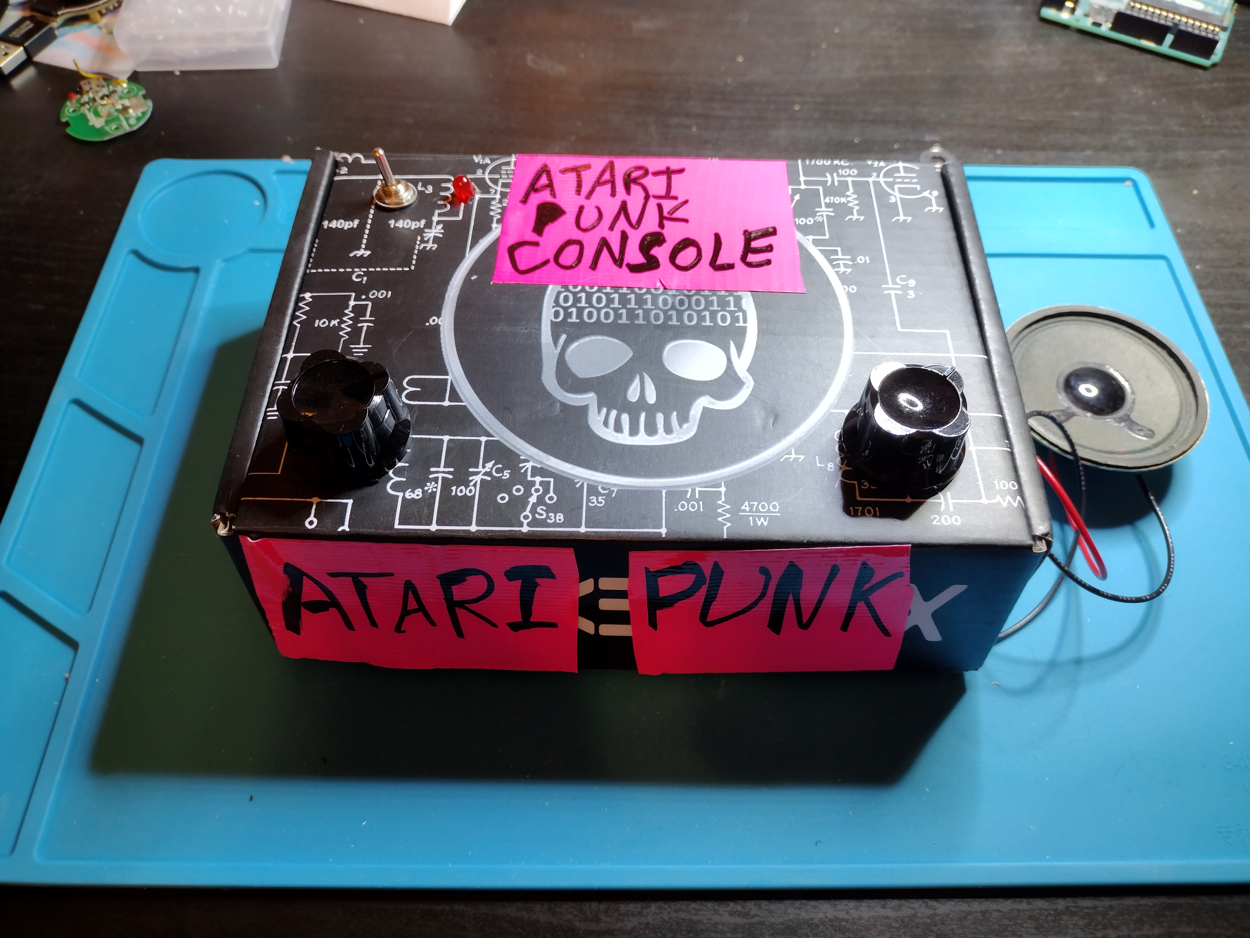 Picture of a black box on a blue anti-static mat. The box has a toggle switch and LED in the upper left corner, two knobs, one in each bottom left corner, and the words "Atari Punk Console" written in sharpie on pink duct tape taped to the top and side of the box