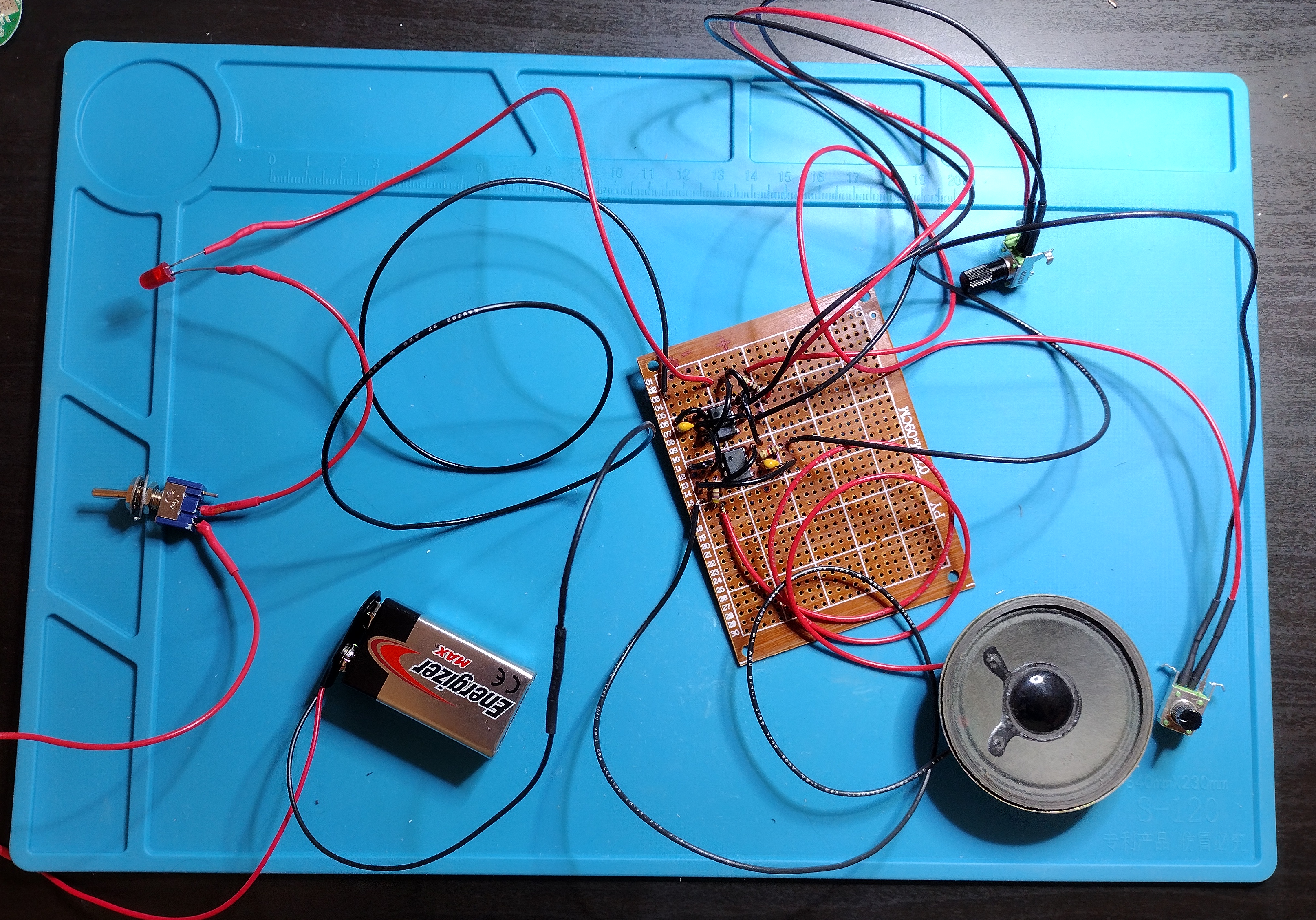 photo of a perfboard on a blue anti-static mat on a black desk. A quarter of the perfboard is full of wires and ICs and resistors. Wires connect the perfboard to a 9V battery holder, a toggle switch, a red LED, 2 poteniometers, and a speaker