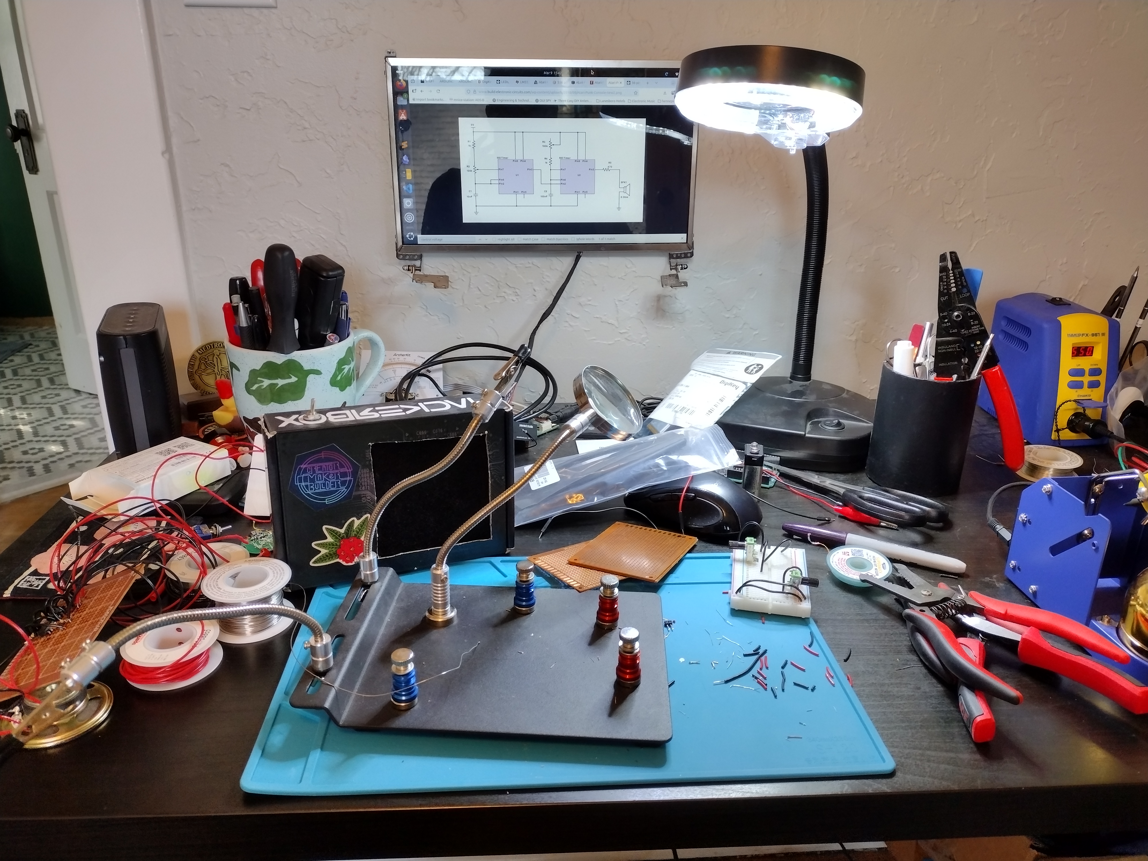 photo of a very cluttered black desk, a blue anti-static mat is in front with a helping hands set for soldering. Wires, hand tools, soldering tools are scattered across the desk. A small monitor can be seend hanging on the wall behind the desk, with a schematic on it.