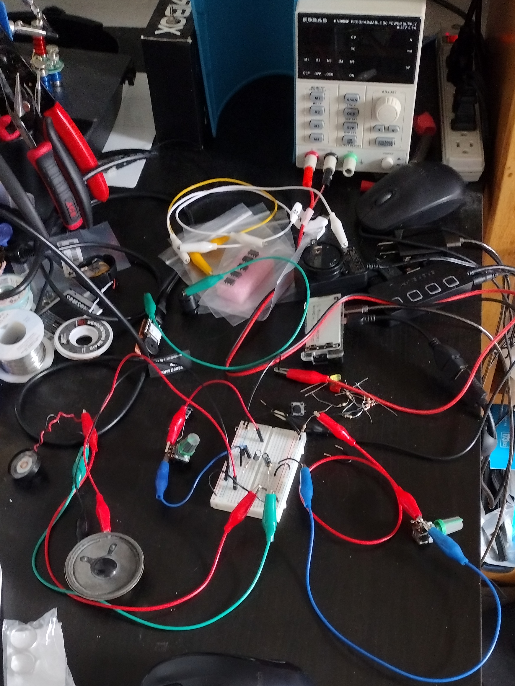 photo of a messy black desk with a breadboard and wires. You can tell the wires are connected to speakers with alligator clips, and there appear to be multiple capacitors, transistors, and resistors on the breadboard, but the exact layout is unclear.