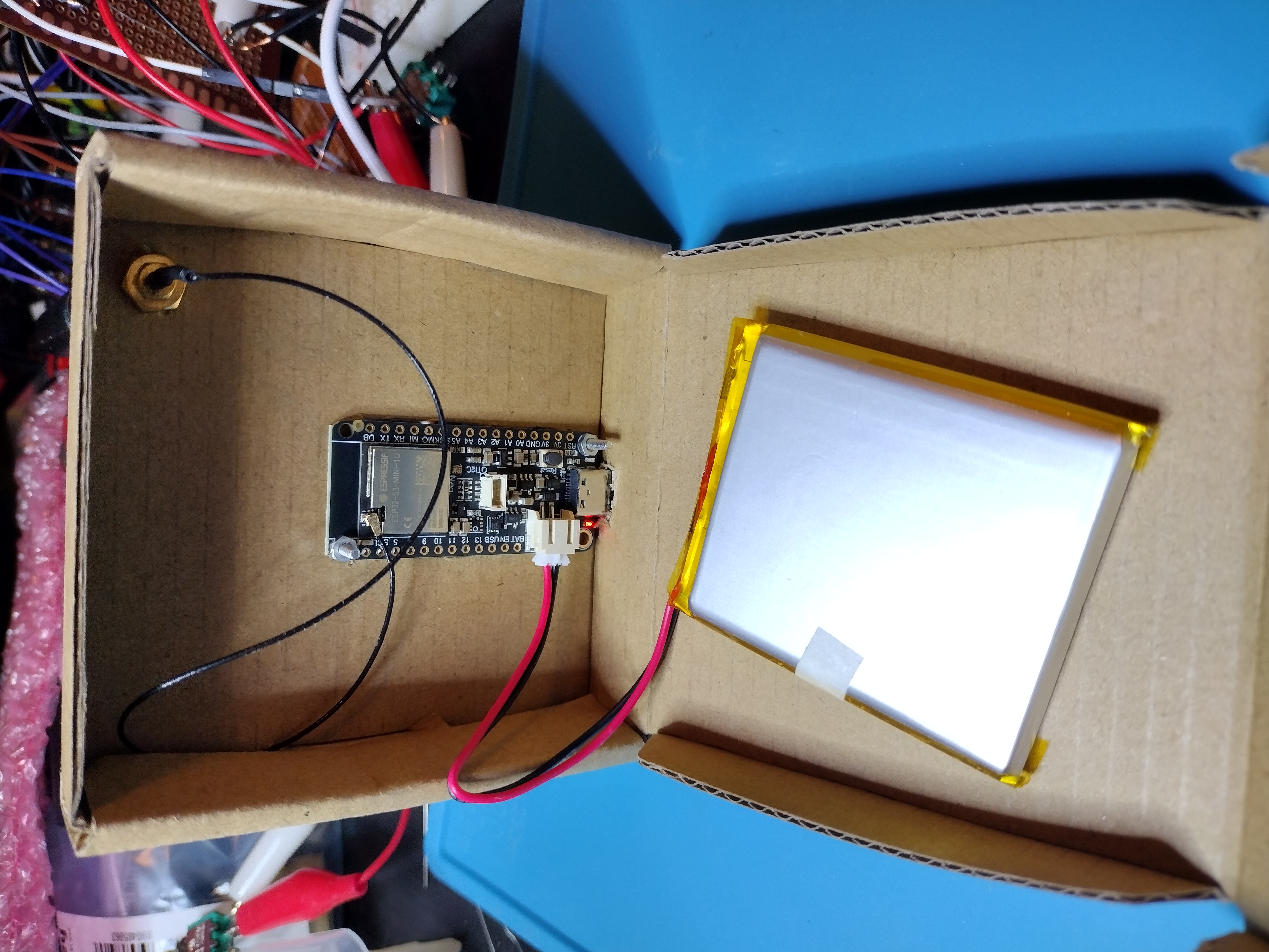 photograph of the inside of a cardboard box. You can see the opposite side of the adafruit feather, and the antenna wire connecting from the feather to the antenna connector outside of the box. There is also a silver lipoly batter with a JST connector plugged into the feather