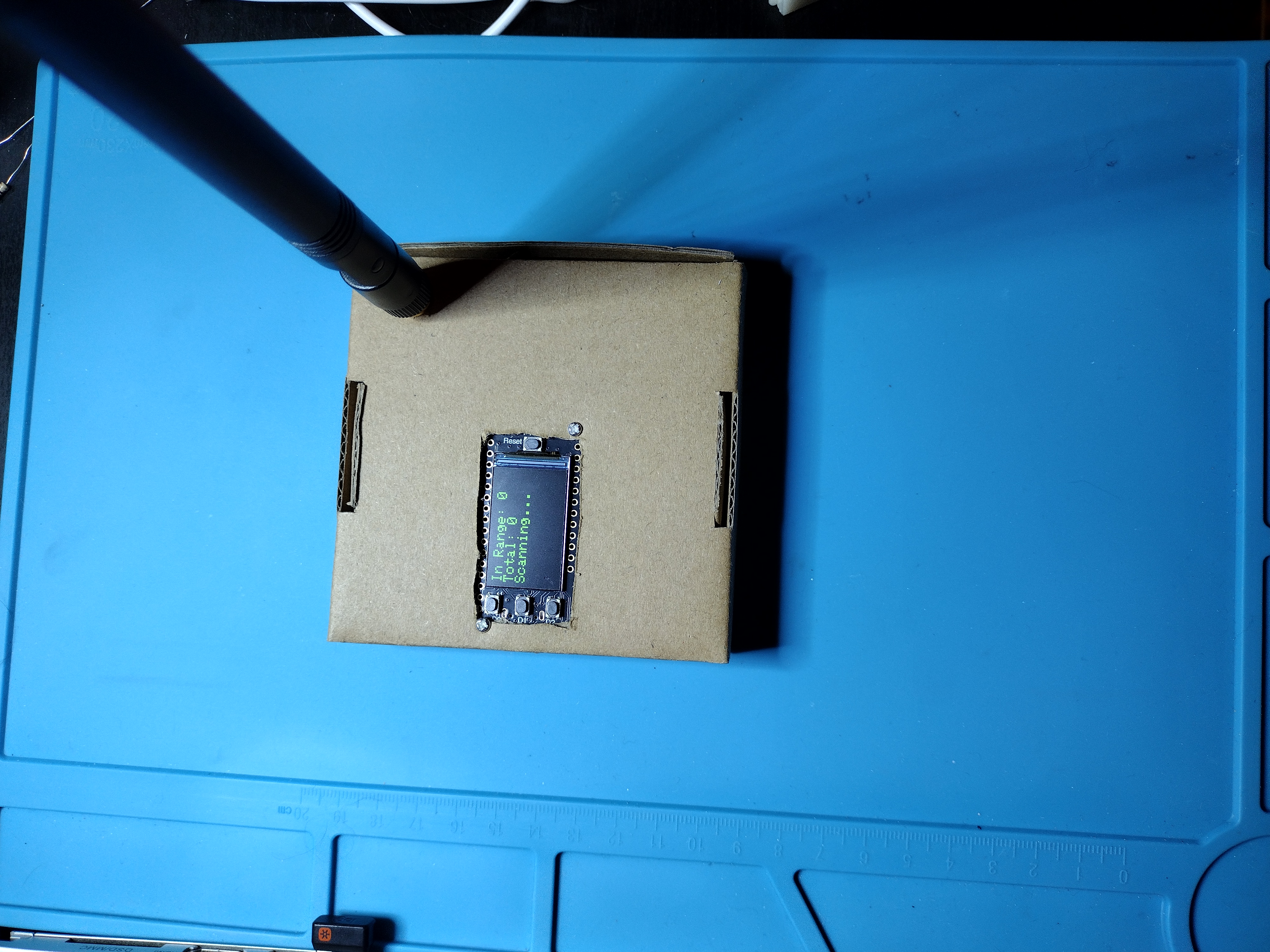 photograph of a small square cardboard box on a blue background. The box has a roughly rectangular hole cut into it, with the adafruit's display showing through the hole. The upper right has a large black omni antenna sticking out. 