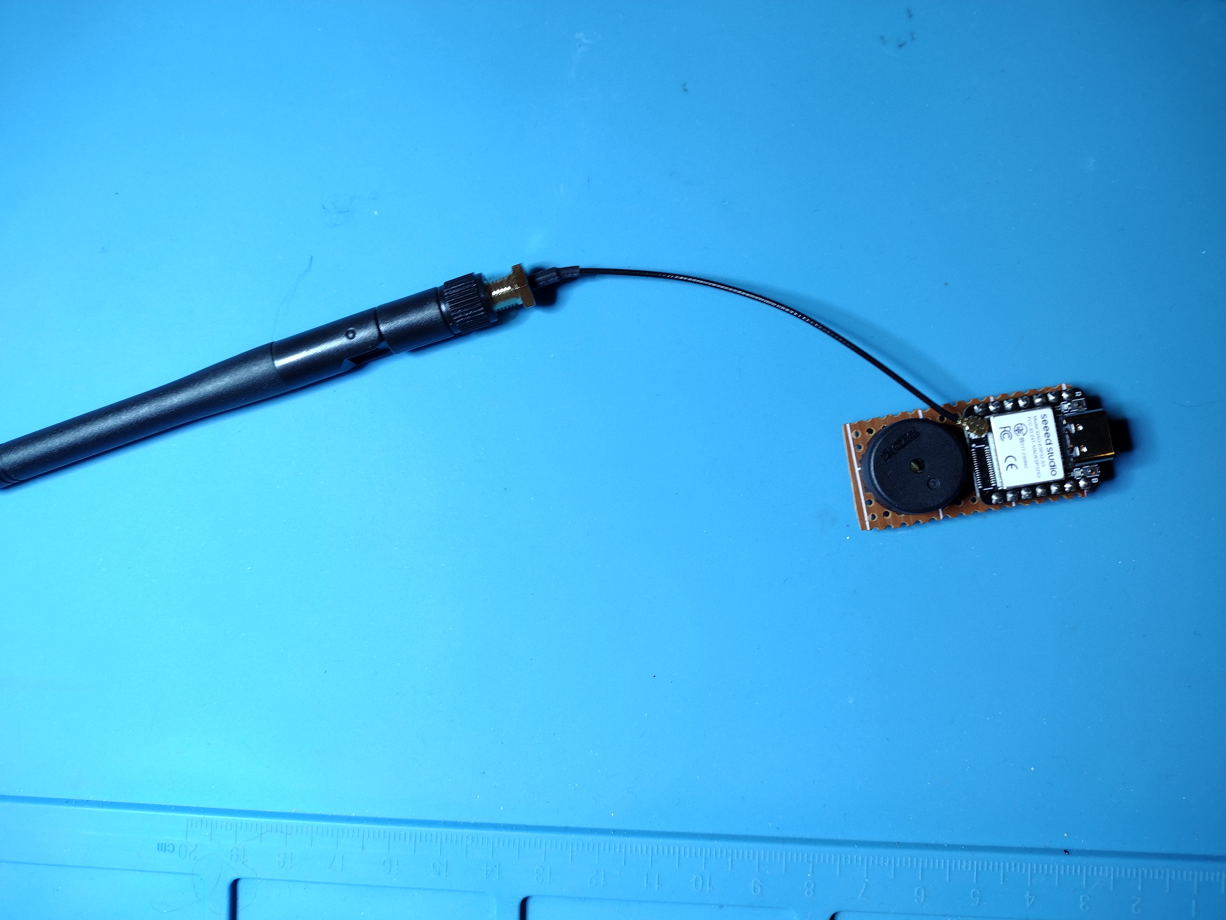 Picture of a large omni-directional antenna connected with a black wire to a small development board, next to a piezo buzzer, on a blue background. You can just barely see that the development board and buzzer are on a brown perf board that has clearly been cut to a small size.