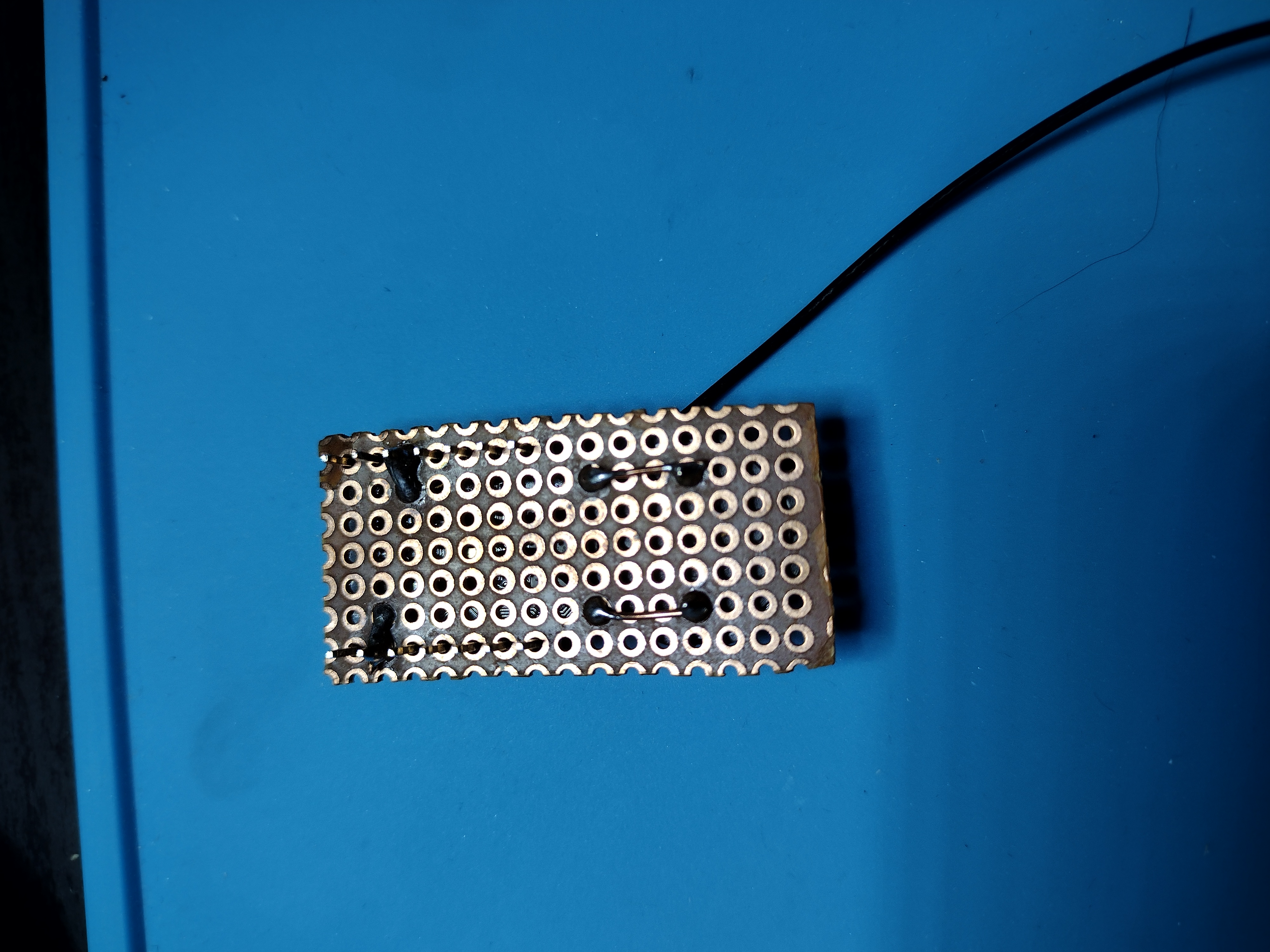 Picture of the backside that is mostly the brown perf board, with edges that are roughly cut to a small size. 2 pins of the development board have been soldered to a hidden wire, which then comes out lower down on the board and connects to pins on the piezo buzzer. The soldering looks slightly blobby but functional.