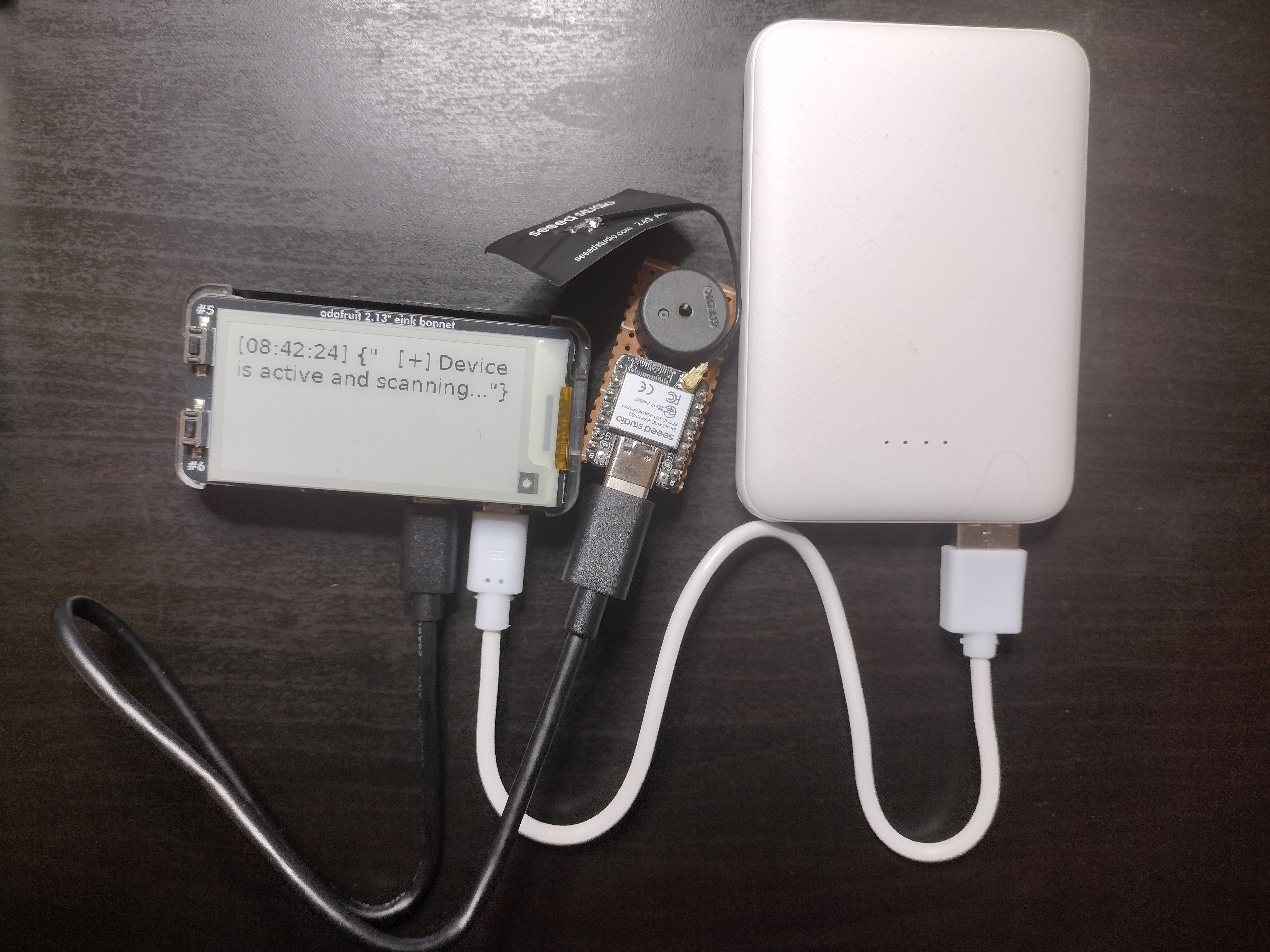 Picture of the raspberry pi with the E-ink screen connected, displaying a timestamp and "Device is active and scanning". It is connected to a white powerbank, and a Xiao ESP32S3 soldered onto perfboard with a piezo buzzer